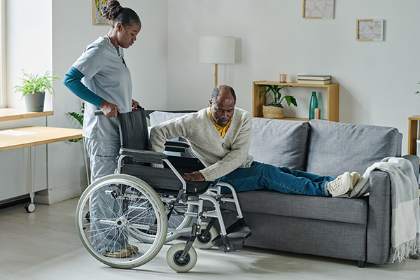 African young social worker helping man with disability to stand up, she caring about him at home